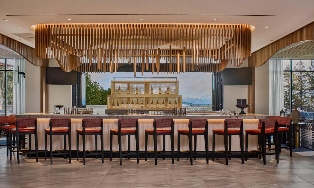 Modern hotel bar at Limelight Mammoth featuring a marble countertop, warm wood ceiling installation, red upholstered bar stools, and mountain views.